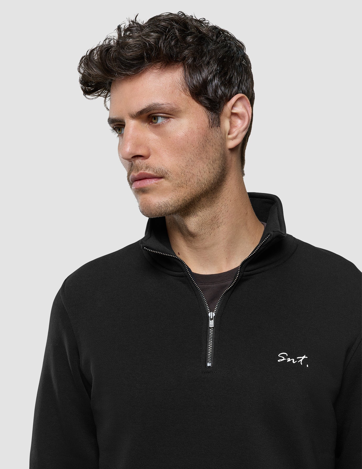 Ein Mann, der ein schwarzes Sweatshirt mit halbem Reißverschluss trägt, auf dessen Brust in weißer Stickerei „Snt.“ steht.